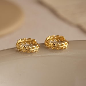 May include: Two gold-colored hoop earrings. Each earring features a detailed leaf design. The earrings are displayed on a light-colored, slightly curved surface, with a soft, blurred background.