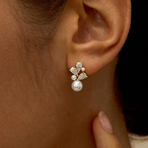 Puede incluir: Primer plano de un pendiente de botón dorado con un diseño de racimo. El pendiente presenta una perla central, rodeada de perlas más pequeñas y cristales claros y brillantes. El pendiente se lleva en una oreja.