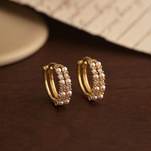 Może przedstawiać: Dwa złote kolczyki koła z potrójnym rzędem. Kolczyki zdobione są małymi, białymi perłami i maleńkimi, przezroczystymi kryształkami. Prezentowane są na ciemnobrązowej powierzchni.