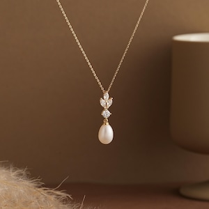 May include: A delicate gold necklace with a pear-shaped pearl pendant. The necklace features a fine chain and is adorned with sparkling, leaf-shaped and round-cut gemstones. The background is a warm, neutral tone, highlighting the jewelry's elegance.
