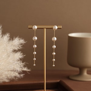 Puede incluir: Elegantes pendientes dorados con una cascada de perlas blancas. Los pendientes se exhiben en un soporte dorado sobre un fondo cálido y neutro. Las perlas varían en tamaño, creando un diseño fluido.