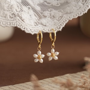 Könnte beinhalten: Ein Paar goldfarbene Creolen mit weißen Blumenanhängern. Jede Blume hat fünf Blütenblätter und eine goldfarbene Mitte. Die Ohrringe hängen vor weißer Spitze und einem verschwommenen braunen Hintergrund.