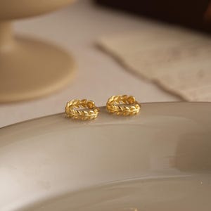 以下が含まれることがあります： 葉のデザインが施された、2つのゴールドカラーのフープピアス。ピアスは小さく丸く、フープの周りに葉の細かい模様があります。明るい色の表面に置かれています。