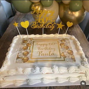 以下が含まれることがあります： 金色と白の装飾が施された白いケーキ。ケーキには「Happy 60th Birthday Paula」と書かれた飾り付けがされています。
