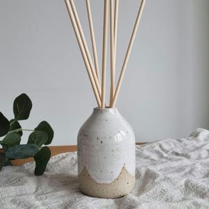 May include: A white ceramic essential oil diffuser with a speckled glaze and a natural, unglazed base. The diffuser holds several wooden reeds, creating a minimalist and natural aesthetic. The diffuser is on a white linen cloth.