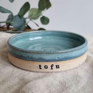 Może przedstawiać: Ręcznie robiona ceramiczna miska z jasno niebieskim szkliwionym wnętrzem i beżowym zewnętrzem. Słowo "tofu" jest wydrukowane na czarno z boku miski. Misa stoi na neutralnej kolorystycznie lnianej powierzchni.