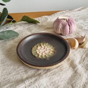 May include: A dark brown ceramic garlic grater with a textured centre, surrounded by a raised rim. A whole purple garlic bulb and several cloves are next to the grater on a neutral linen surface. Kitchenware for culinary use.