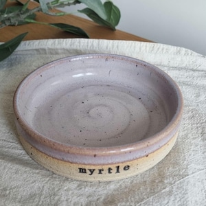 May include: A handmade ceramic bowl with a light purple interior and a tan exterior. The word "myrtle" is printed in black on the side of the bowl. The bowl is round and has a slightly raised rim.