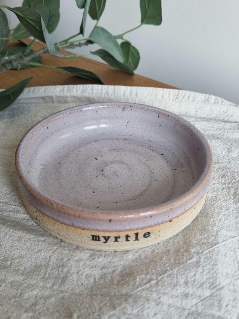 May include: A handmade ceramic bowl with a light purple interior and a tan exterior. The word "myrtle" is printed in black on the side of the bowl. The bowl is round and has a slightly raised rim.
