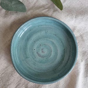 Może przedstawiać: Ręcznie robiony, turkusowy ceramiczny talerz o lekko teksturowanej powierzchni. Talerz ma płytki, zaokrąglony kształt z ciemniejszą krawędzią i spiralnym wzorem pośrodku. Talerz jest prawdopodobnie używany do serwowania potraw.