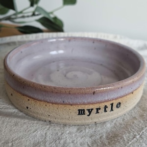 May include: A handmade ceramic bowl with a lavender interior and a tan exterior. The rim is a darker shade of lavender. The word "myrtle" is printed in black on the side of the bowl. The bowl is sitting on a white surface.
