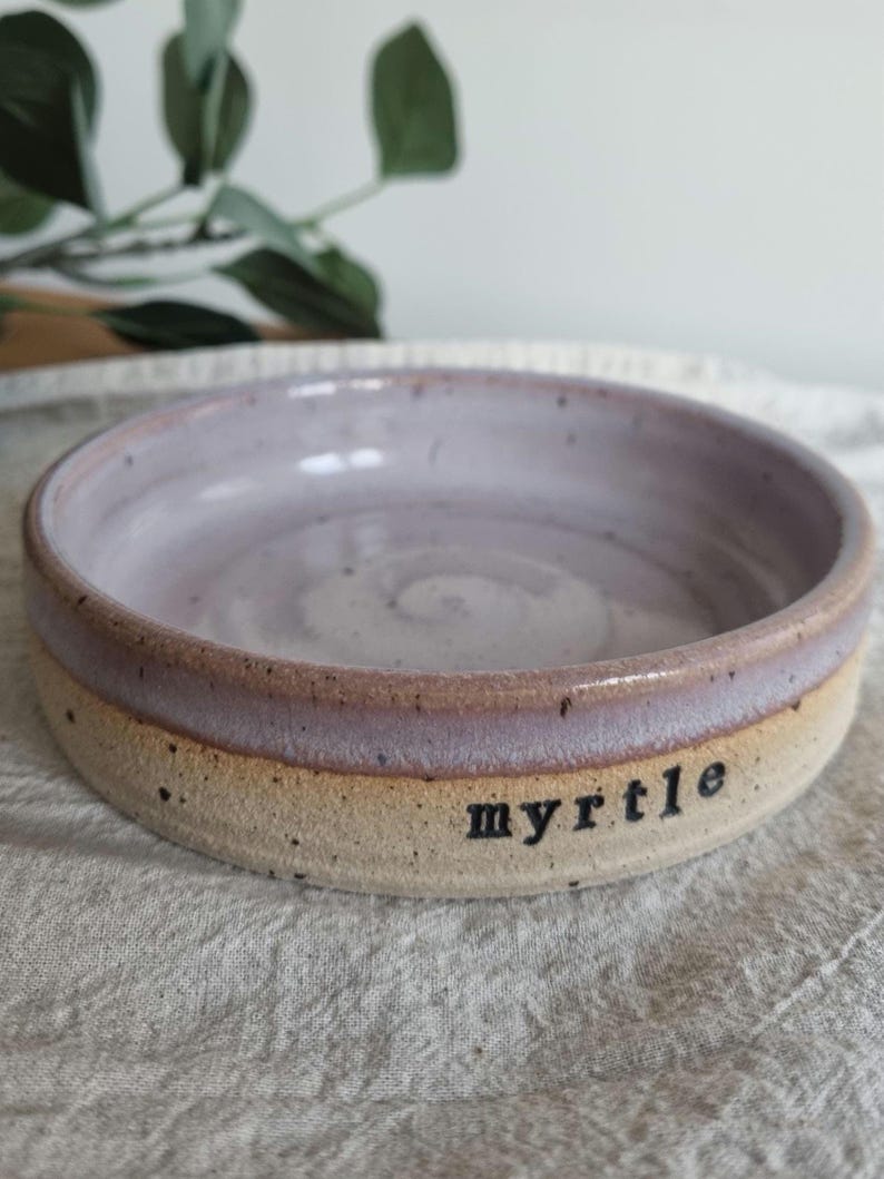 May include: A handmade ceramic bowl with a lavender interior and a tan exterior. The rim is a darker shade of lavender. The word "myrtle" is printed in black on the side of the bowl. The bowl is sitting on a white surface.