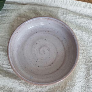 May include: A handmade ceramic bowl with a speckled, light pink interior and a darker pink rim. The bowl has a shallow design and sits on a textured, off-white surface. The bowl's diameter is approximately 6 inches.