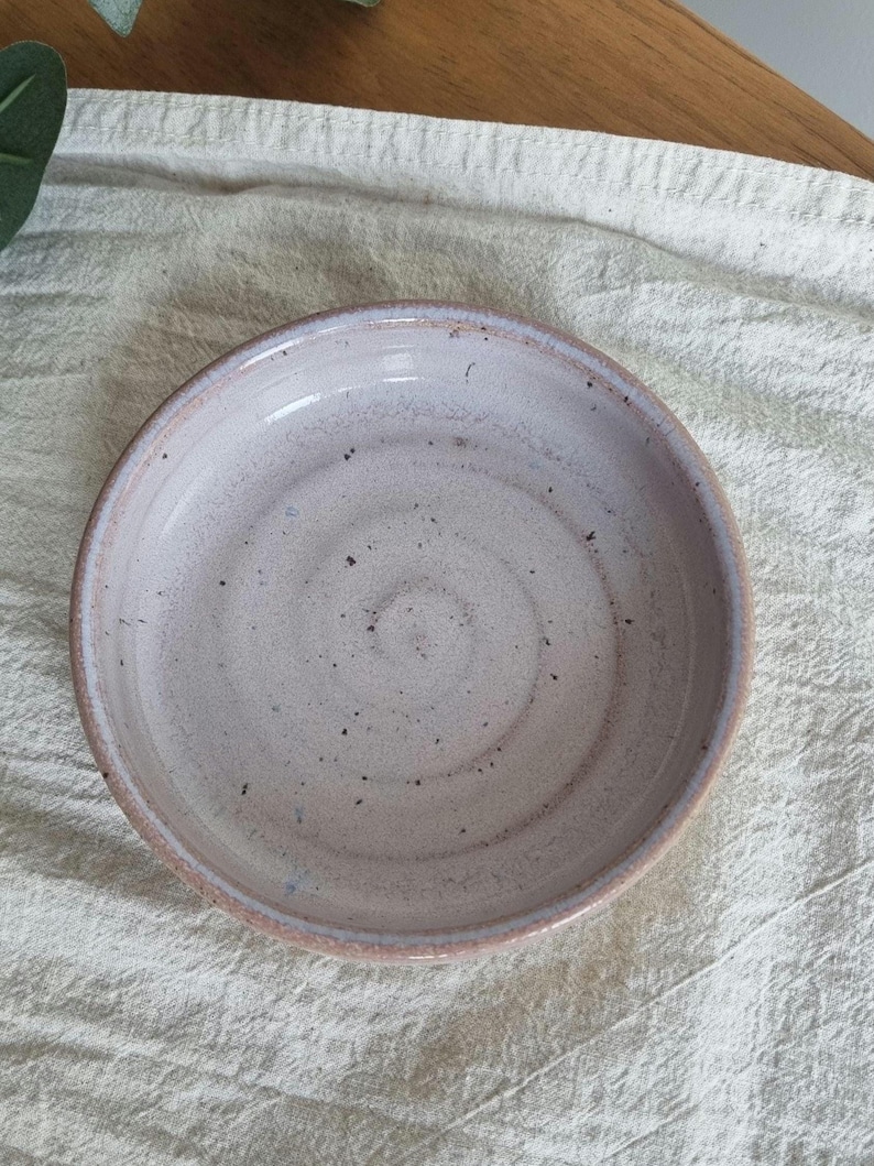 May include: A handmade ceramic bowl with a speckled, light pink interior and a darker pink rim. The bowl has a shallow design and sits on a textured, off-white surface. The bowl's diameter is approximately 6 inches.