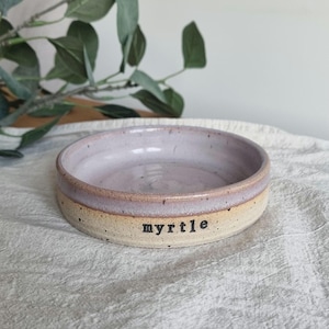 May include: A handmade ceramic bowl with a lavender interior and a tan exterior. The word "myrtle" is printed in black on the exterior. The bowl is round and sits on a white cloth.