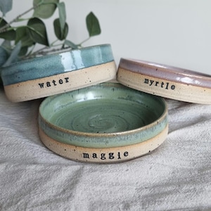 May include: Three handmade ceramic pet bowls in shades of blue, green, and purple. Each bowl has a natural, unglazed base and a glazed interior. The bowls are labeled with the names "water", "myrtle", and "maggie".