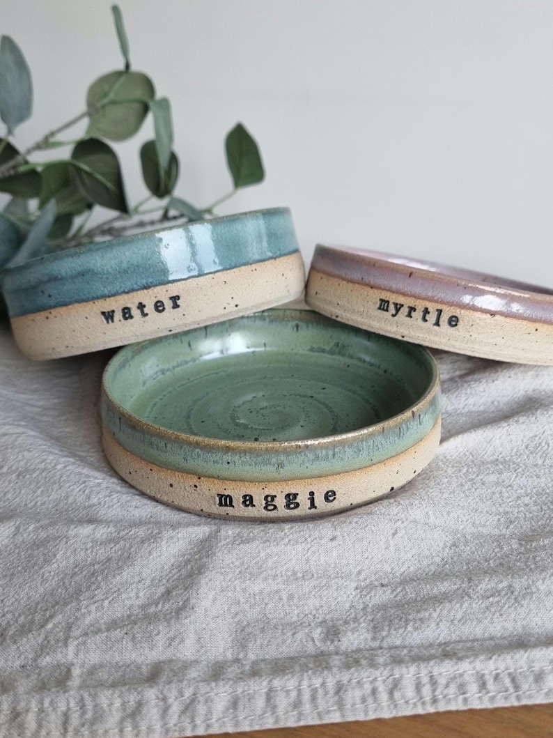 May include: Three handmade ceramic pet bowls in shades of blue, green, and purple. Each bowl has a natural, unglazed base and a glazed interior. The bowls are labeled with the names "water", "myrtle", and "maggie".
