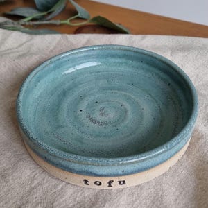 Może przedstawiać: Ręcznie robiona ceramiczna miska z turkusowym wnętrzem i beżowym zewnętrzem. Słowo "tofu" jest wydrukowane na czarno z boku miski. Miska jest okrągła i ma niewielką głębokość, odpowiednią do serwowania małych porcji.