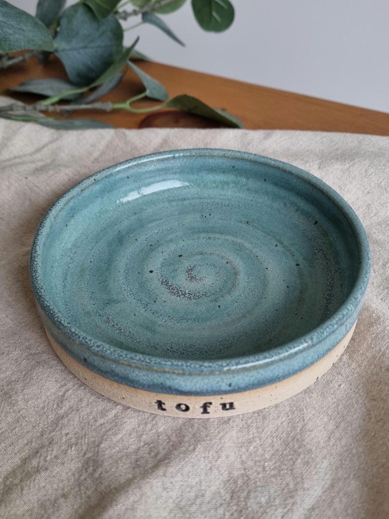 Może przedstawiać: Ręcznie robiona ceramiczna miska z turkusowym wnętrzem i beżowym zewnętrzem. Słowo "tofu" jest wydrukowane na czarno z boku miski. Miska jest okrągła i ma niewielką głębokość, odpowiednią do serwowania małych porcji.