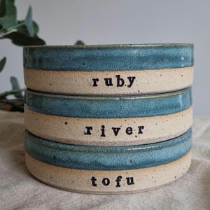 Może przedstawiać: Ułożone w stos ceramiczne miski z turkusową obw&oacute;dką i beżową podstawą. Każda miska ma czarny napis: "ruby", "river" i "tofu". Miski są ułożone jedna na drugiej.