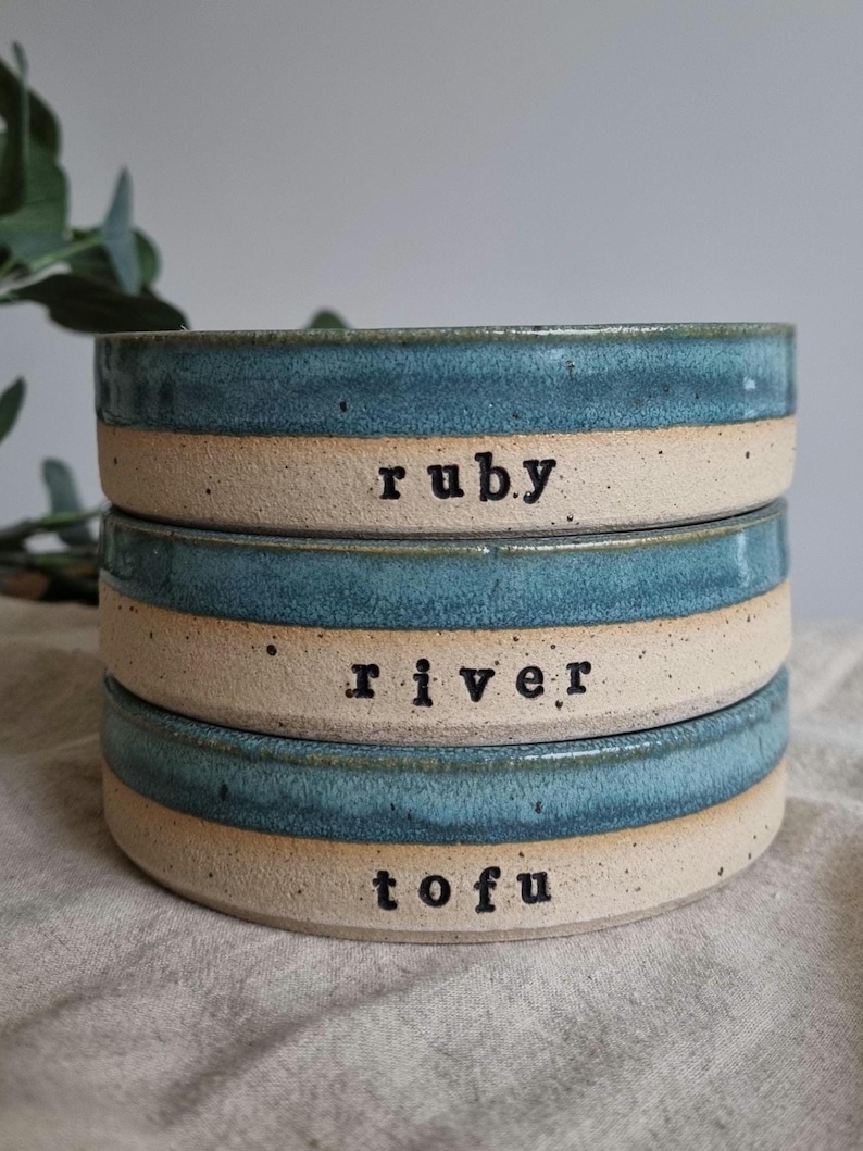 Może przedstawiać: Ułożone w stos ceramiczne miski z turkusową obw&oacute;dką i beżową podstawą. Każda miska ma czarny napis: "ruby", "river" i "tofu". Miski są ułożone jedna na drugiej.