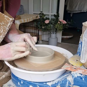 Może przedstawiać: Osoba pracuje na kole garncarskim, formując jasną ceramiczną miskę. Koło jest biało-niebieskie, widoczne są narzędzia i glina. Osoba nosi rękawice i fartuch, skupiona na rzemiośle.