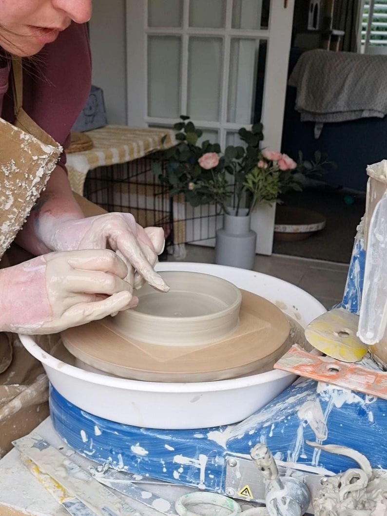 Może przedstawiać: Osoba pracuje na kole garncarskim, formując jasną ceramiczną miskę. Koło jest biało-niebieskie, widoczne są narzędzia i glina. Osoba nosi rękawice i fartuch, skupiona na rzemiośle.