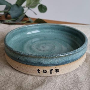 Może przedstawiać: Ręcznie robiona ceramiczna miska z turkusowym wnętrzem i beżowym zewnętrzem. Słowo "tofu" jest wydrukowane na czarno z boku miski. Miska jest okrągła i wydaje się być glazurowana.