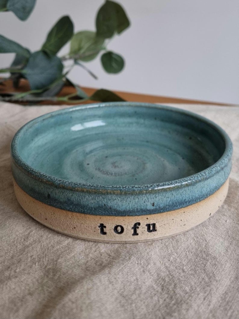 Może przedstawiać: Ręcznie robiona ceramiczna miska z turkusowym wnętrzem i beżowym zewnętrzem. Słowo "tofu" jest wydrukowane na czarno z boku miski. Miska jest okrągła i wydaje się być glazurowana.
