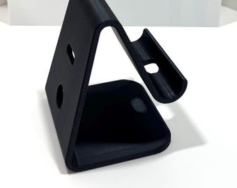 May include: A black, angled phone stand with two circular holes and a curved top. The stand is made of a matte material. Above the stand is a red sign that reads "@3DesignsUSA".