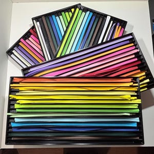 May include: A collection of colorful paper strips arranged in black trays. The strips are organized by color, ranging from red, orange, yellow, green, blue, and purple. The trays are stacked, creating a vibrant display of craft supplies.