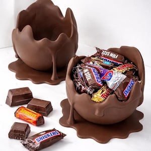 May include: Two chocolate egg-shaped bowls with a melted chocolate design. One bowl is filled with assorted candy bars, including Snickers and Twix. Several unwrapped chocolate pieces and candy bars are scattered around the bowls.