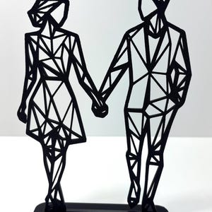 May include: Black metal sculpture of a couple holding hands. The figures are stylized with geometric shapes, standing on a rectangular base. The sculpture is a modern, minimalist design.