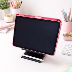 May include: A pink tablet case with a black screen sits on a black stand, with a white stylus in front. Other desk accessories, such as pencils, a keyboard, and a small plant, are visible in the background.