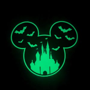 May include: A glowing green silhouette of Mickey Mouse ears with a spooky castle and bats. The design is set against a black background, creating a Halloween theme. The castle has dripping details, and bats are flying around the ears.