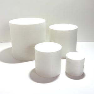 May include: Four white cylindrical display stands of varying heights. The stands are made of a smooth, solid material and are arranged on a white surface. The cylinders are ideal for showcasing products in a retail or studio setting.