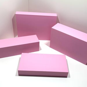 Puede incluir: Cuatro bloques rectangulares rosas de diferentes tamaños están dispuestos sobre una superficie blanca. Los bloques son de color rosa sólido y tienen un acabado mate. La disposición sugiere una exhibición o presentación de producto.