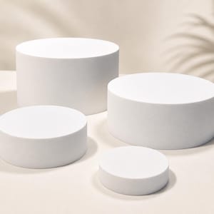 Pode incluir: Quatro suportes de exibição cilíndricos brancos de diferentes alturas e diâmetros estão dispostos sobre uma superfície bege clara. Os suportes são sólidos e lisos, proporcionando uma estética limpa e minimalista. Uma sombra de folha de palmeira é projetada no fundo.
