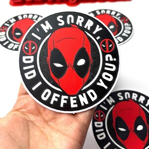 May include: A round black and white sticker with a red Deadpool character in the center. The sticker says "I'm sorry. Did I offend you?" in white text.