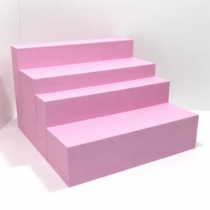 Puede incluir: Un expositor rosa de cuatro niveles. El expositor está construido con bloques rectangulares, creando una plataforma escalonada. El color es un rosa suave y pastel, y el expositor está sobre un fondo blanco. El diseño es simple y moderno, adecuado para exhibir productos.