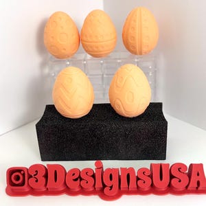 Può includere: Cinque uova di Pasqua color pesca con diversi motivi in rilievo. Le uova sono disposte su una superficie di schiuma nera con un cartello di plastica rosso che recita "@3DesignsUSA".