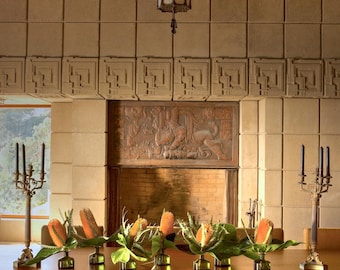 May include: A dining room with a large wooden table set with floral arrangements. The room features a decorative chandelier, a fireplace with a relief carving, and matching candle holders. The chairs have red wooden frames and beige fabric.