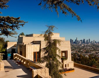 May include: Exterior view of a beige, multi-level building with a patterned facade, set against a clear blue sky. The building overlooks a cityscape with tall buildings. Trees frame the view.