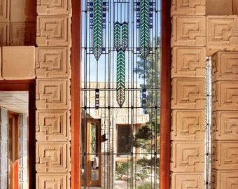 May include: A tall, rectangular stained-glass window with a geometric design, framed by a wooden border. The window features blue and green accents against a clear background, set within a sandstone wall with a repeating square pattern. The window overlooks a garden.