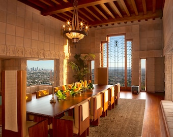 May include: A spacious dining room with a long wooden table set for a meal. The room features large windows offering a city view, a decorative chandelier, and wooden chairs. The walls are made of textured stone.