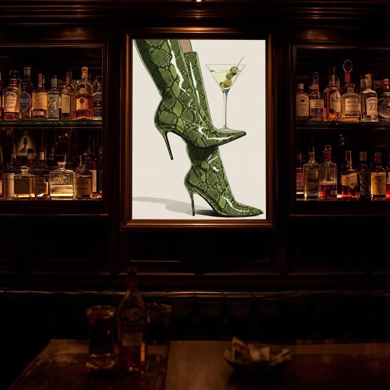 Op de afbeelding: Ingelijste kunst met groene, slangenleer-patroon hoge hakken en een martini glas met olijven. De afbeelding bevindt zich in een bar, met planken vol flessen op de achtergrond.