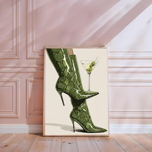 Op de afbeelding: Een ingelijste print met groene laarzen met slangenprint en een martini glas met olijven. Het kunstwerk staat tegen een neutrale achtergrond. De laarzen zijn in een dynamische pose geplaatst.