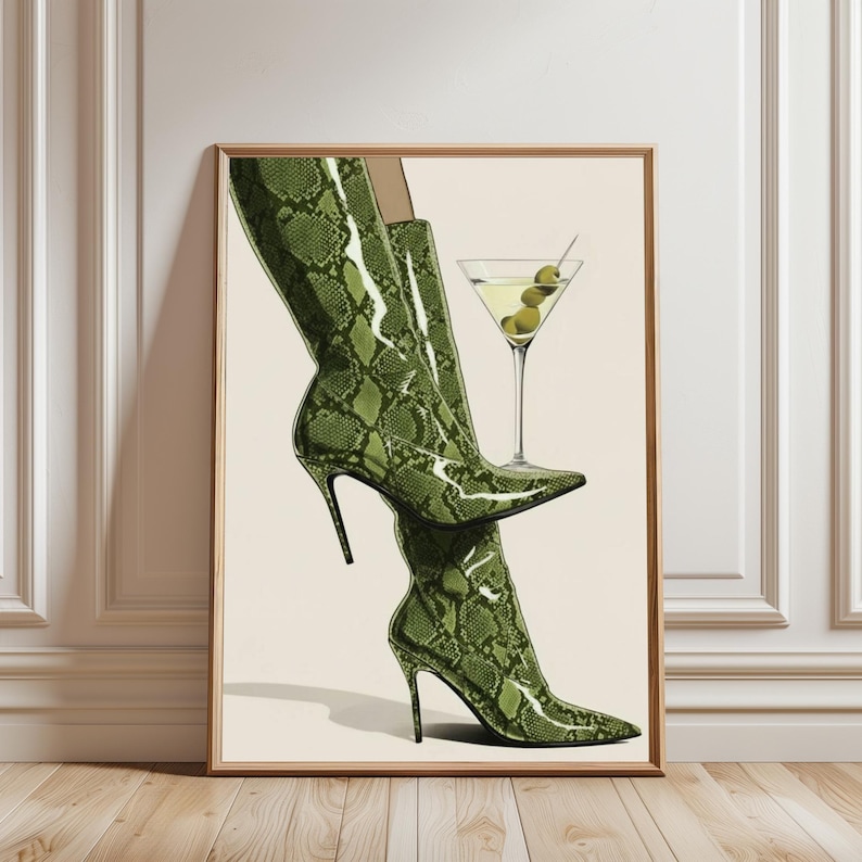 Op de afbeelding: Een illustratie van groene laarzen met slangenpatroon en hoge hakken met spitse tenen. Een martini glas met olijven staat naast de laarzen. Het kunstwerk is ingelijst in een houten lijst en leunt tegen een witte muur.