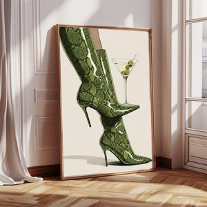 Op de afbeelding: Een ingelijste kunstdruk met een paar groene laarzen met slangenprint en een martini-glas met olijven. Het kunstwerk heeft een neutrale achtergrond en wordt getoond in een kamer met een houten vloer.