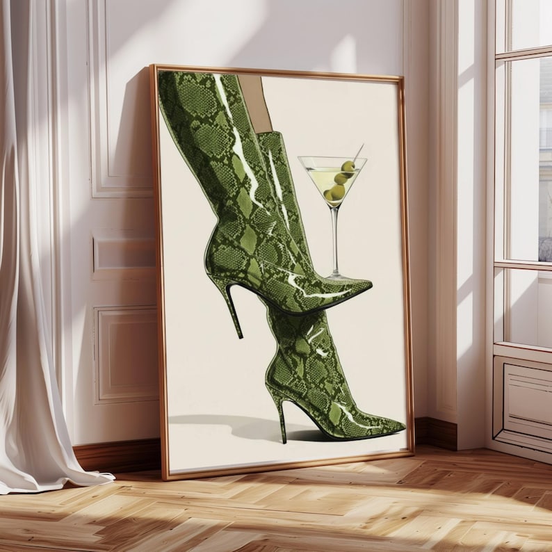Op de afbeelding: Een ingelijste kunstdruk met een paar groene laarzen met slangenprint en een martini-glas met olijven. Het kunstwerk heeft een neutrale achtergrond en wordt getoond in een kamer met een houten vloer.